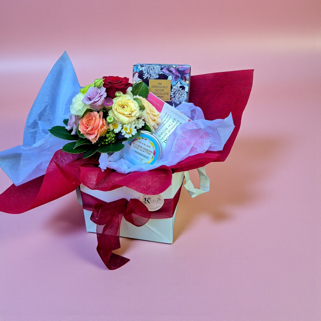 Bloom & Treat gift box containing a posie of seasonal flowers, bar of chocolate, Botanical Bath Bomb and flower seed scatter by Bee & Bloom, wrapped in red paper and presented in a white gift box, tied with a matching ribbon.