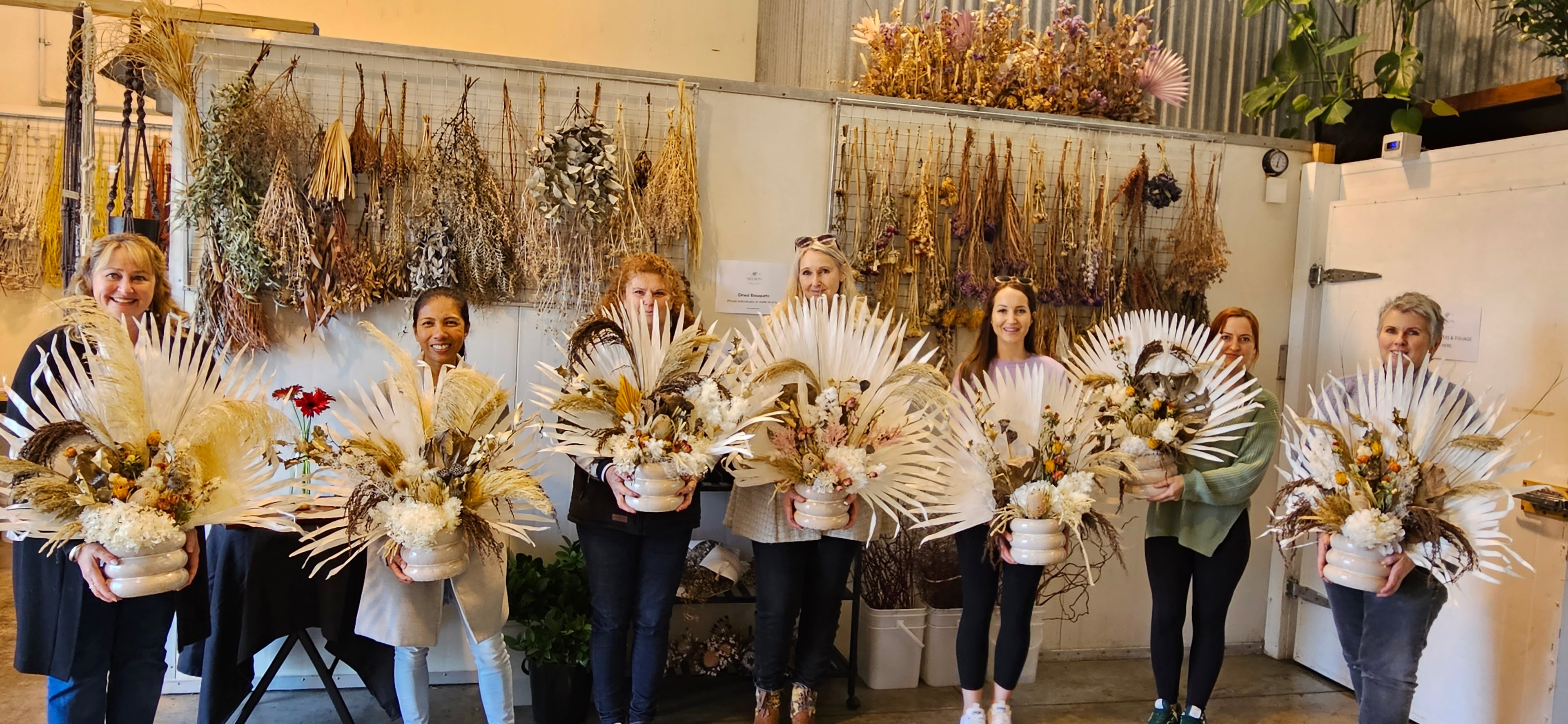 A group of women smiling and holding up their Dried Floral Arrangements which they made in Nelson Flower Company's workshop.
