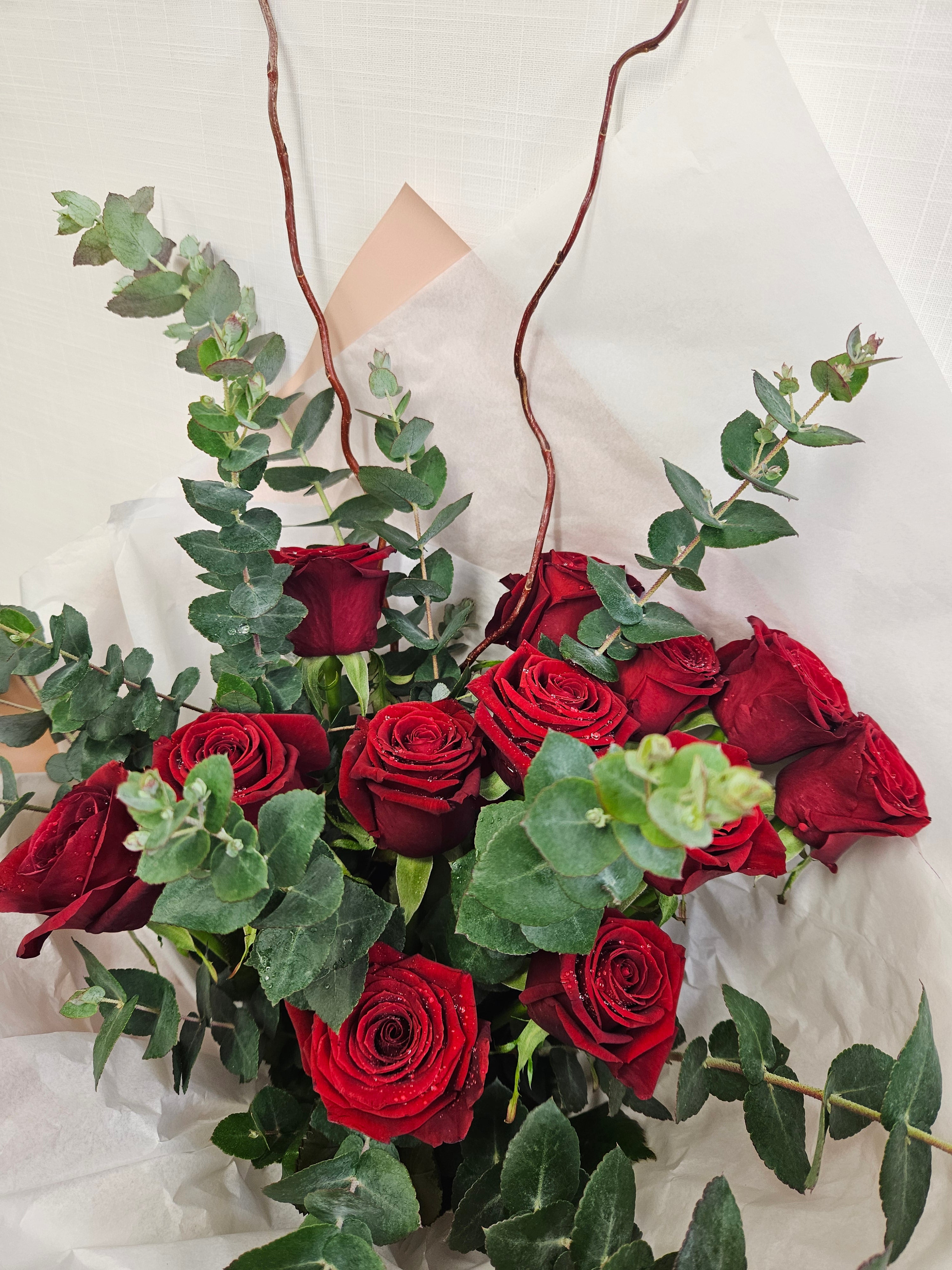 12 Reasons bouquet of vibrant red roses and foliage, wrapped in champagne and white tissue paper.
