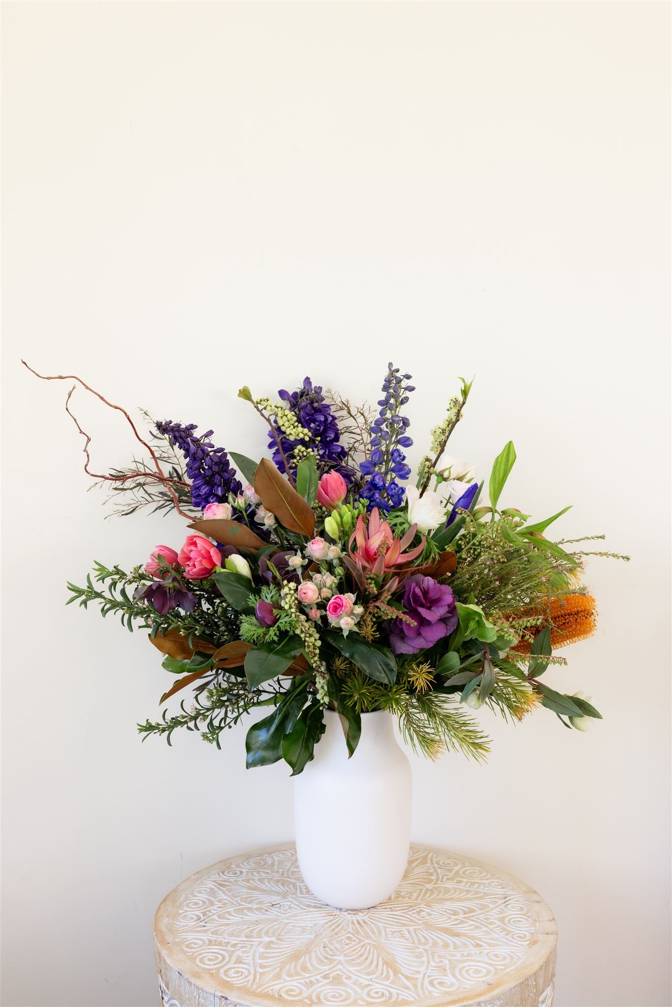 Florist Choice Divine Bunch bouquet made with purple and pink flowers with foliage, presented in a white vase atop a carved wooden table (presentation only).