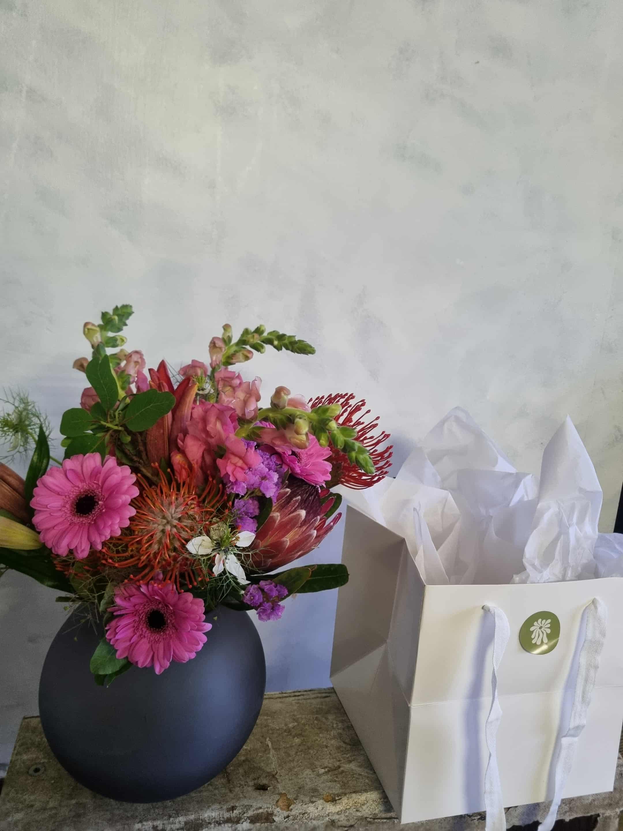 Bowl of Brights arrangement made with vibrant flowers and foliage, presented in a fishbowl vase and packaged in a white gift bag. Choose an optional add-on of chocolate or a candle.