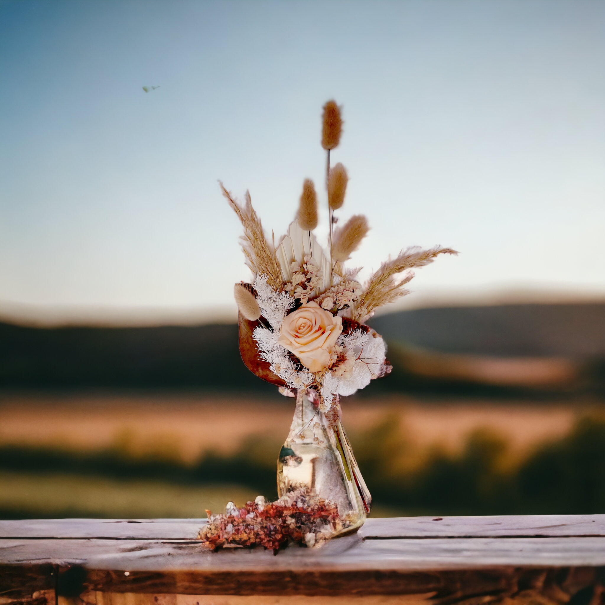 Eternal Petals bouquet with neutral-toned flowers and foliage, arranged in a glass vase on a bench at sunset overlooking the countryside.