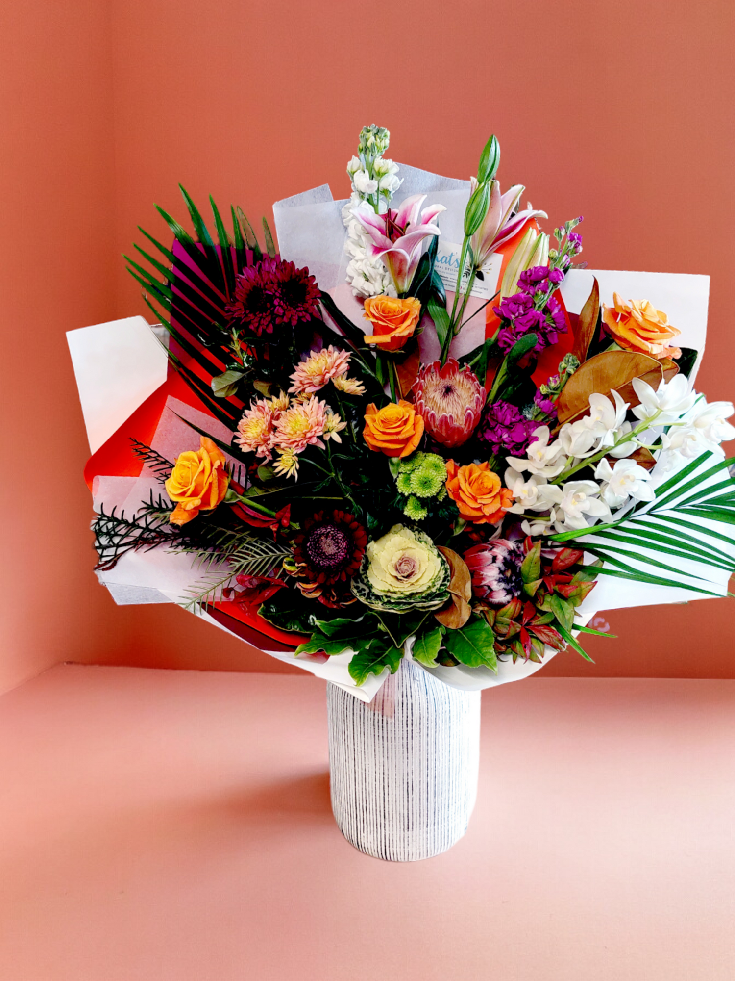 Sympathy Flowers bouquet in Large size made with warm-toned flowers and foliage, wrapped in white and purple paper.