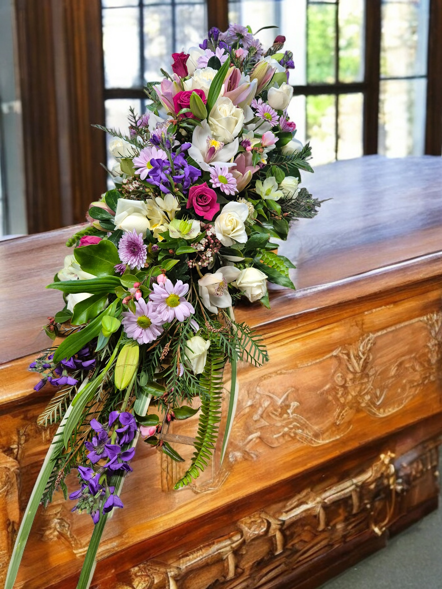 Bright Casket Spray for funerals and memorials made with an assortment of brightly coloured purple, pink and white flowers and foliage.