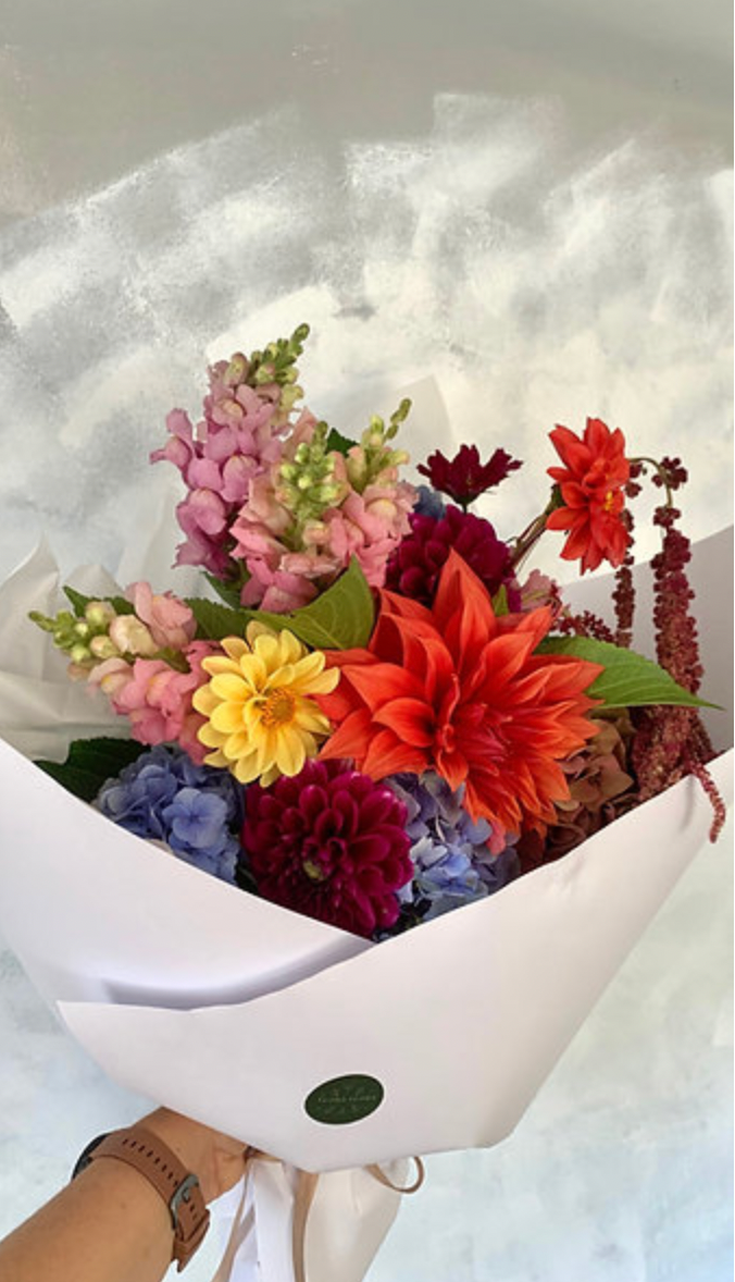 Bright Delight bouquet made with a variety of vibrant seasonal flowers in warm tones, wrapped in white paper tied with a ribbon.