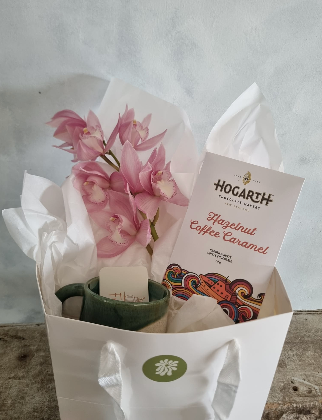 Orchid Giftbox with a beautiful pink orchid bloom, bar of Hogarth Chocolate in Hazelnut Coffee Caramel, and a handmade green mug by Francis Pottery, presented in a white gift bag.