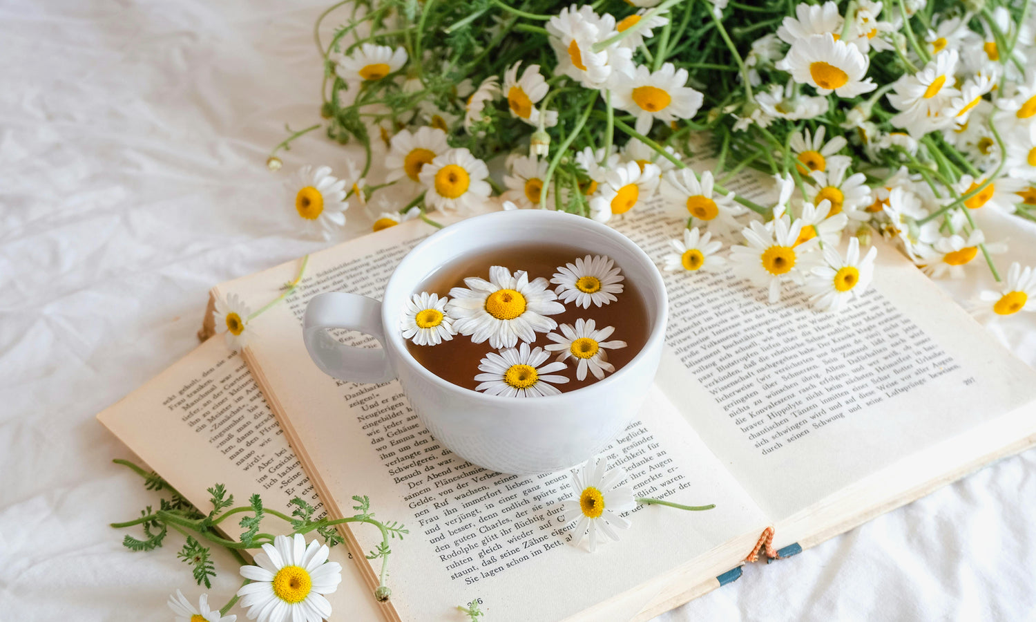 Cup of chamomile tea made from fresh flowers rests atop a pile of open books next to a bouquet. Photo by hello aesthe on Unsplash.