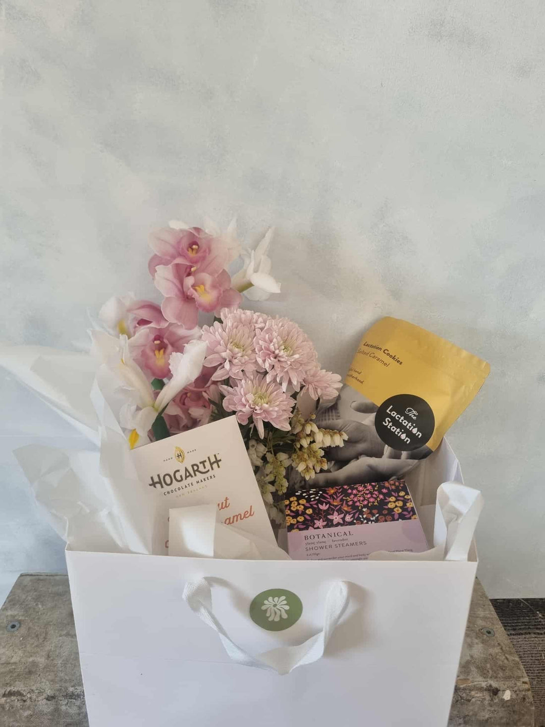 Hey Mama Gift Box complete with soft pastel floral bouquet in ceramic pedestal vase, Lactation Station Salted Caramel Cookies, a box of shower steamers, and a bar of Hogarth Chocolate, all presented in a white gift bag.