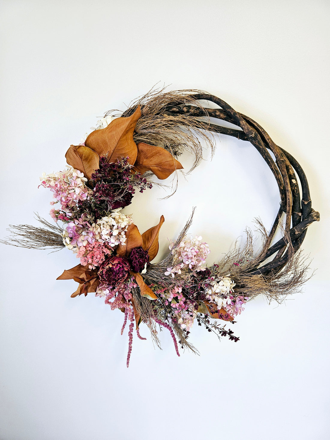 Dried floral wreath with pink, red, white flowers and neutral brown foliage hanging on a wall