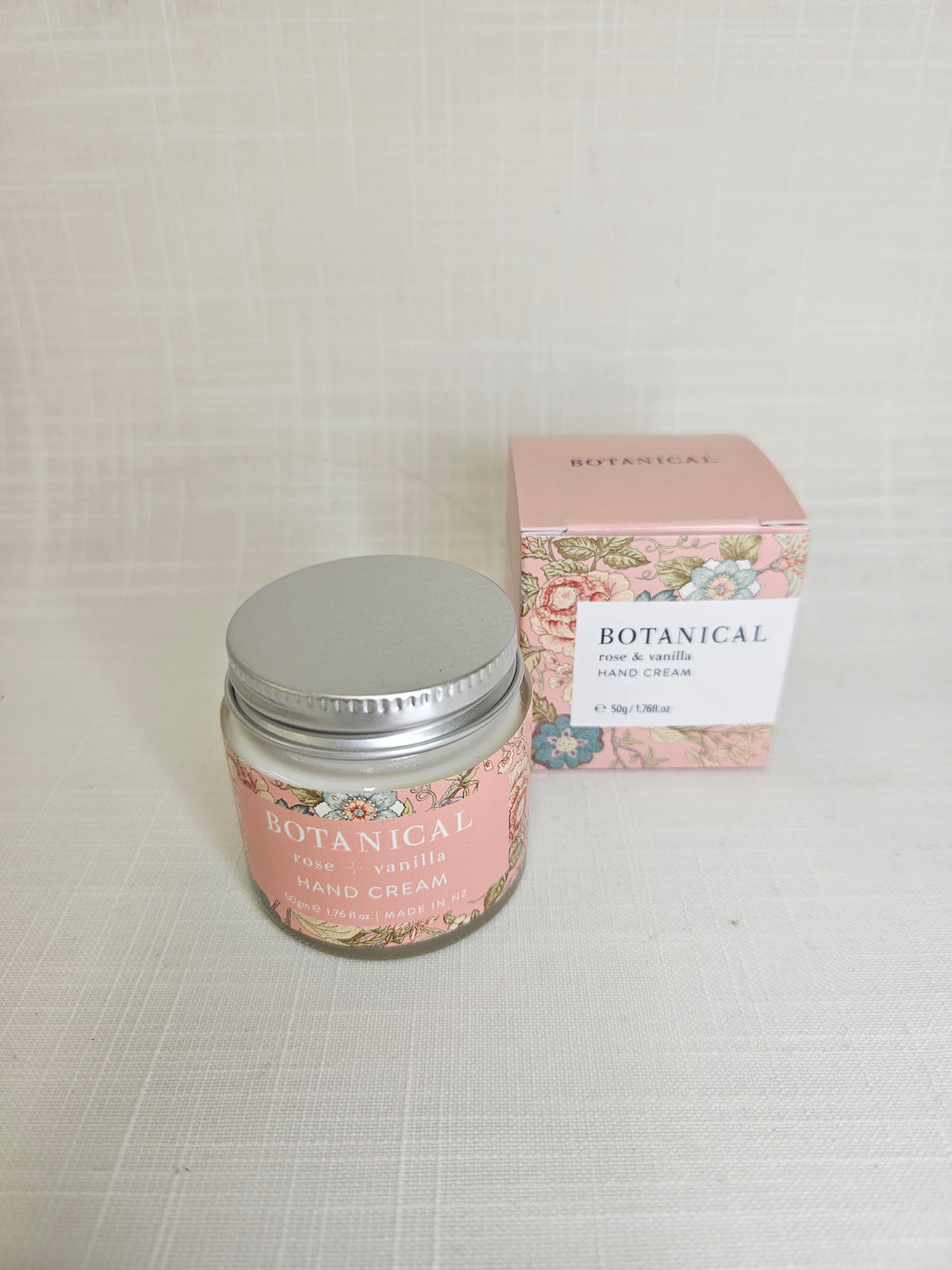 A jar of the Botanical Rose &amp; Vanilla handcream with a pink floral label stands next to a matching box.