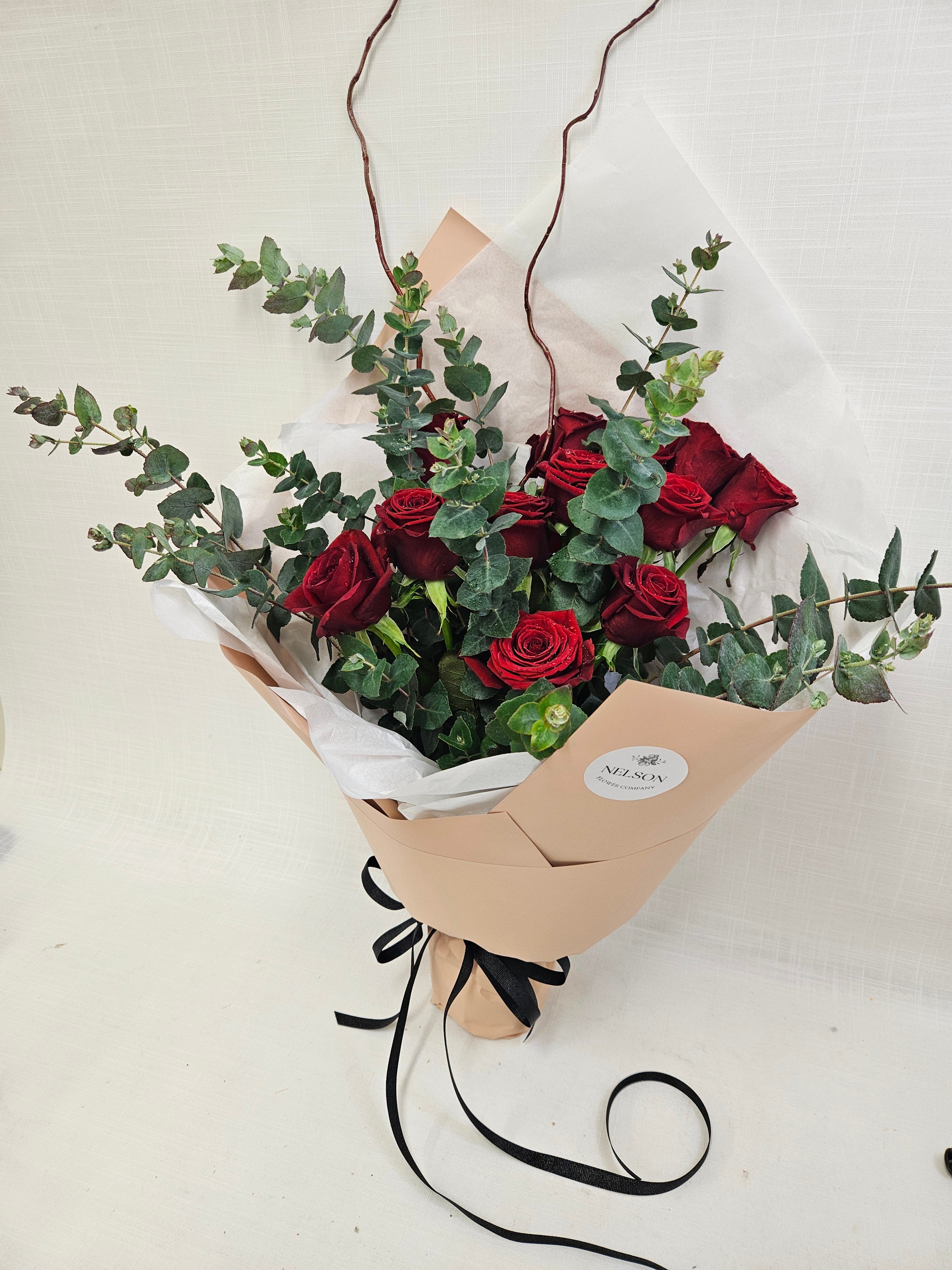 12 Reasons bouquet of vibrant red roses and foliage, wrapped in champagne and white tissue paper.