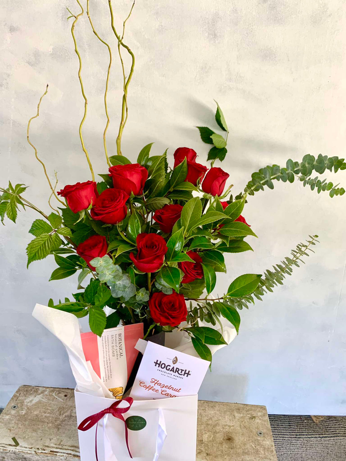 The Ultimate gift basket complete with a ceramic vase of a dozen red roses, chocolate and a bath bomb, presented in a white gift bag.