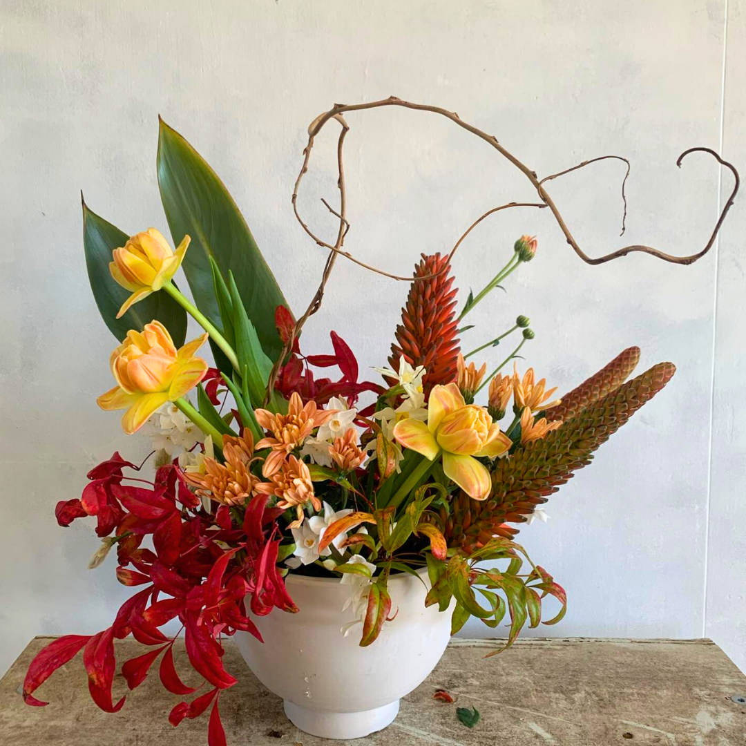 Botanical Bedlam floral arrangement with red, orange and yellow flowers and foliage, presented in a white vase.