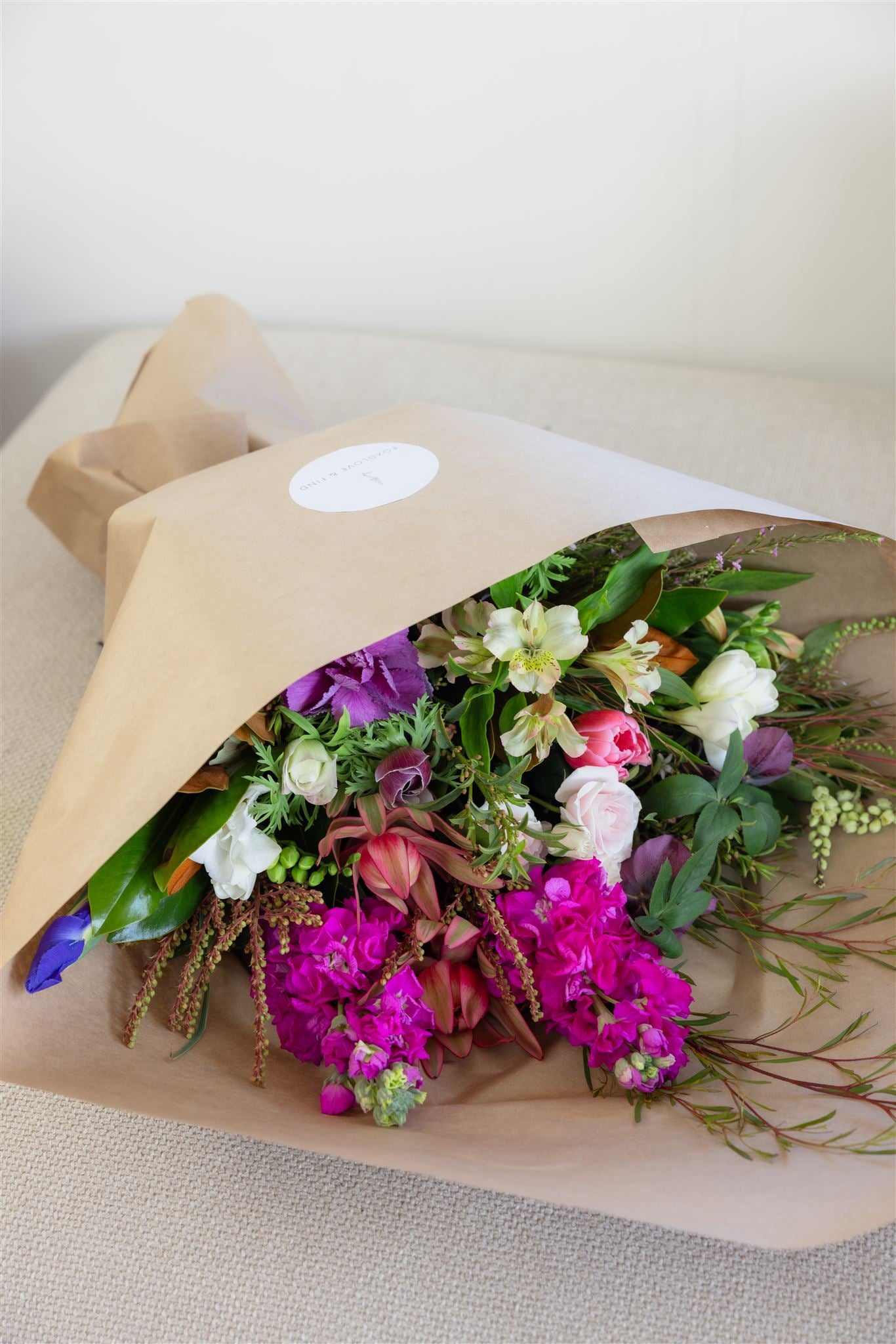 Florist Choice bouquet made with purple and hot pink flowers with foliage, wrapped in brown paper, secured with a sticker and ribbon.