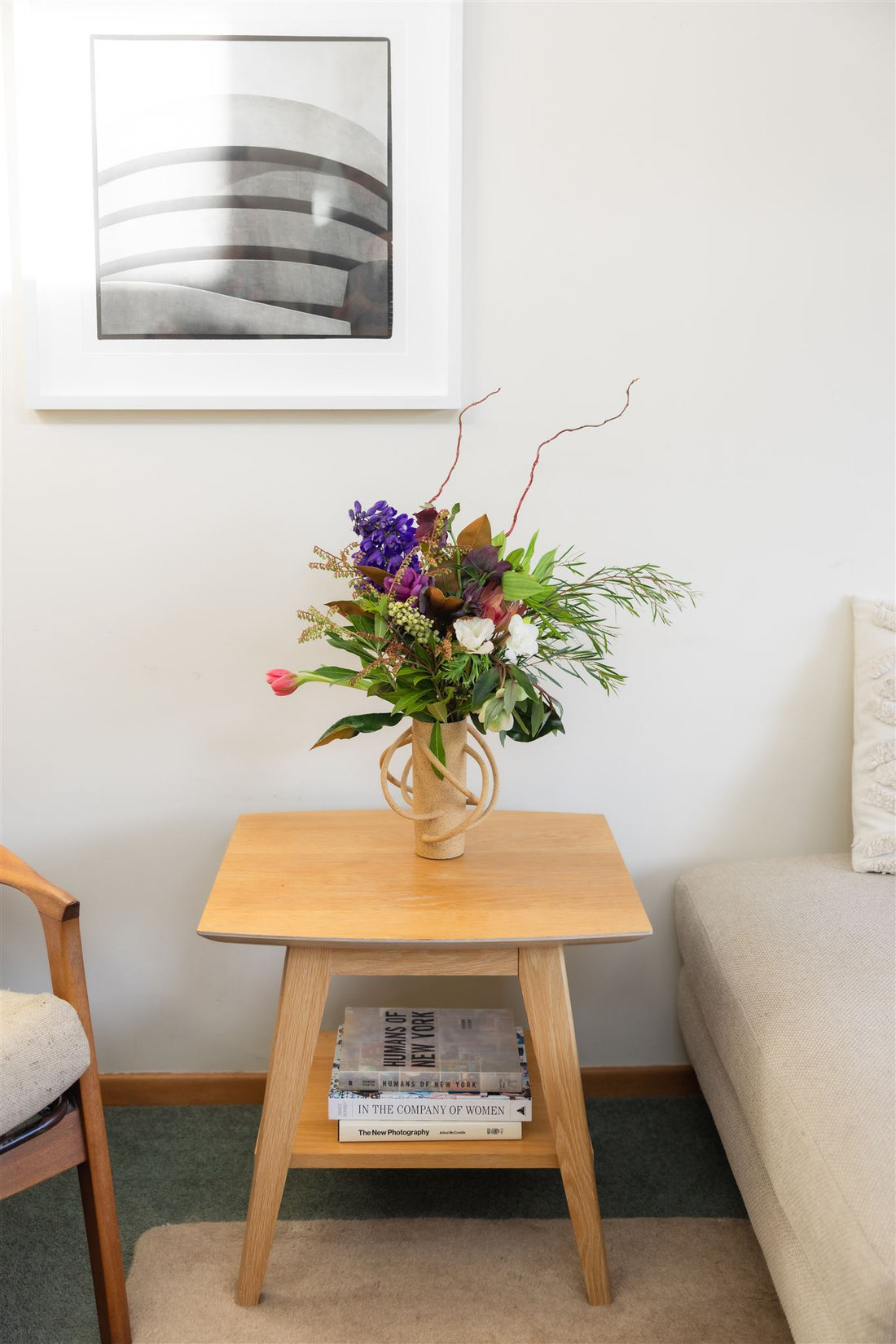 Florist Choice Divine Bunch bouquet made with purple and pink flowers with foliage, presented in an organic-designed vase atop a bedside table (presentation only).