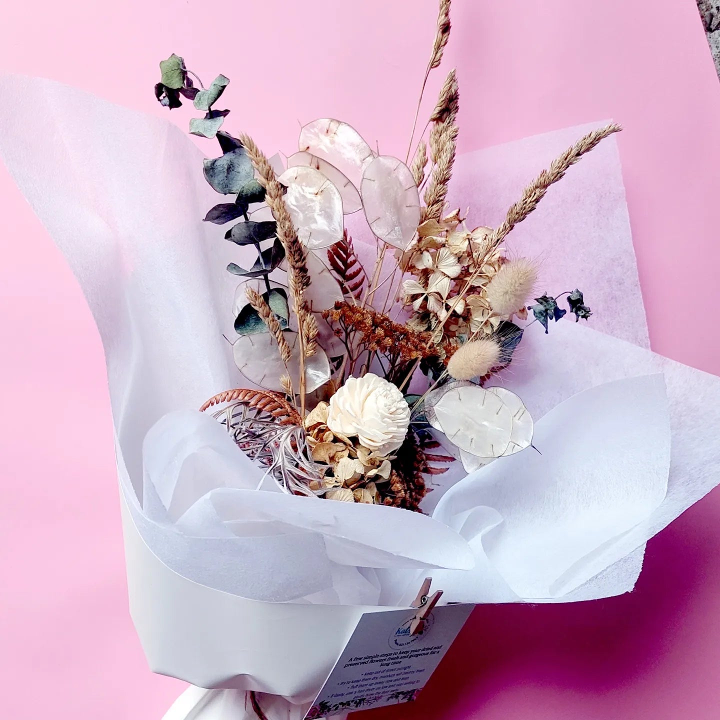 Eternal Petals bouquet with neutral-toned flowers and foliage, wrapped in white paper against a pink backdrop.