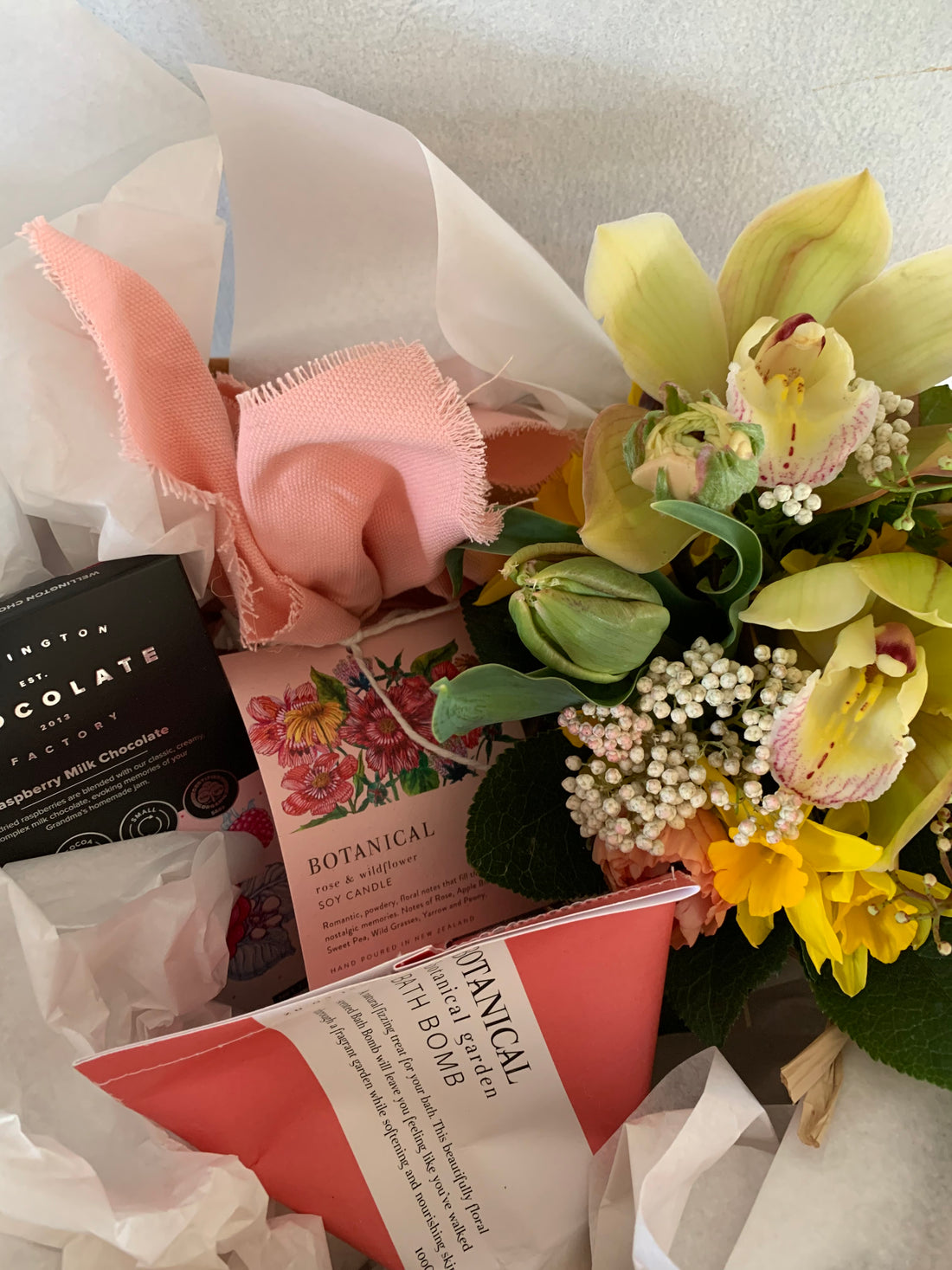 Seasonal Posie Gift Box comprised of a small variety of flowers, chocolate, bath bomb and a candle, packaged in a white gift bag with matching tissue paper and ribbon.