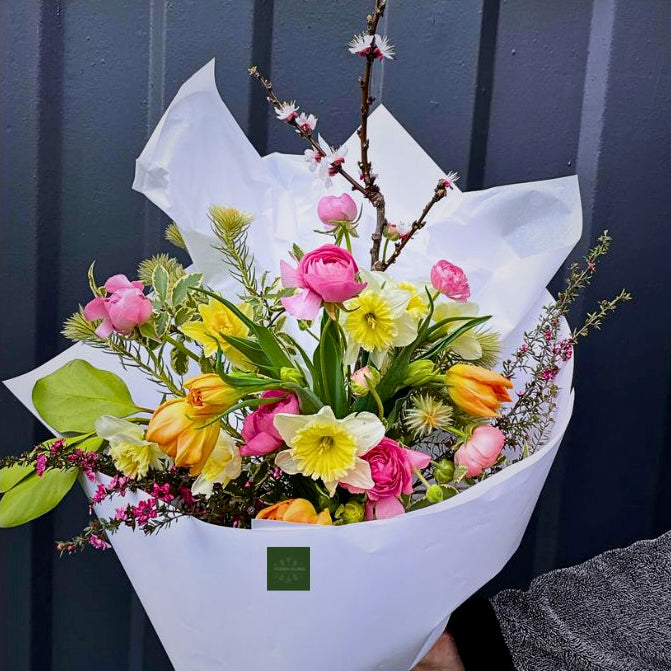 An example of what your Seasonal Bunch from Flora Flora could look like. Bright springtime flowers in yellow, orange and pink, with green foliage wrapped in white tissue paper.