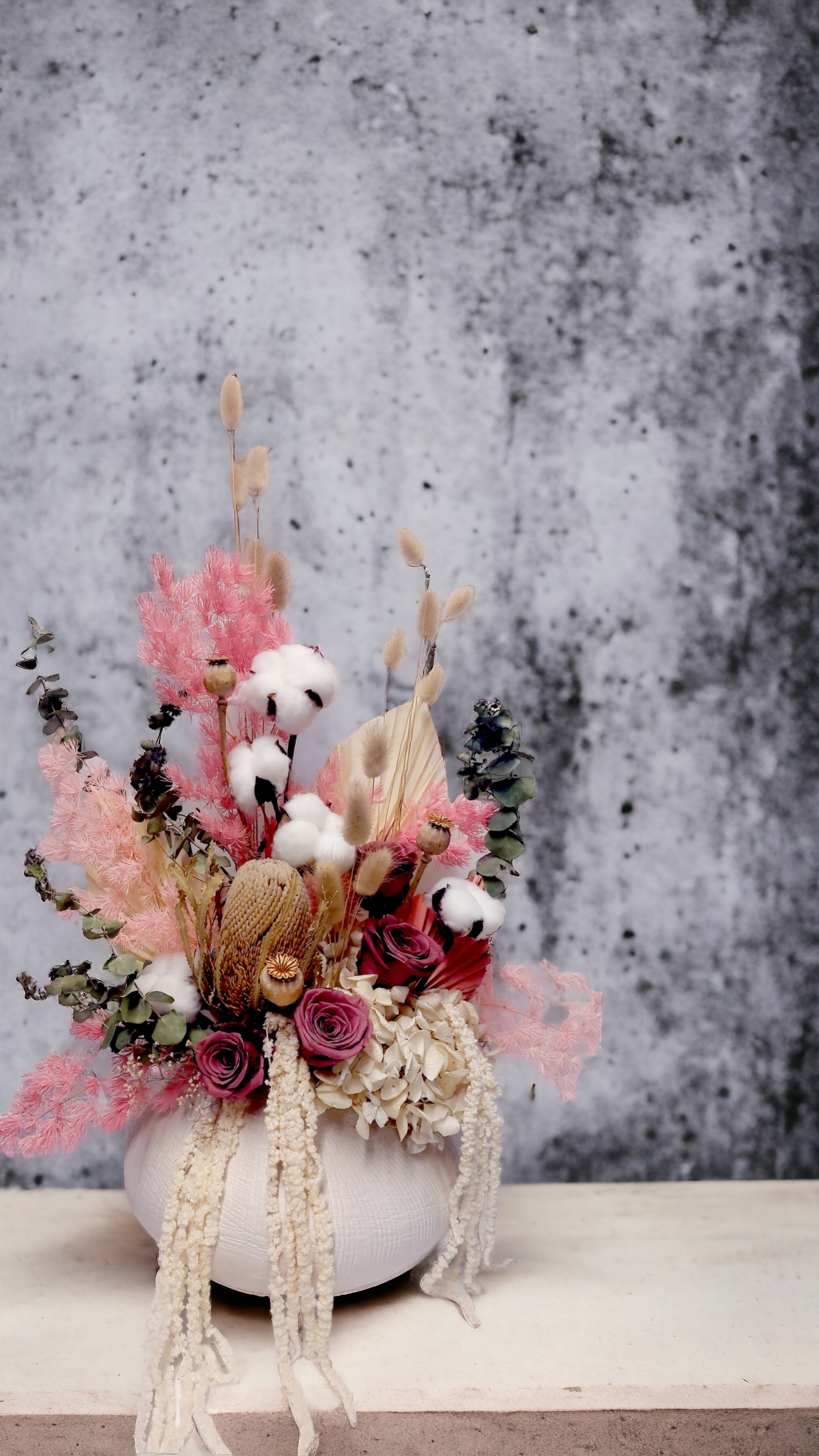 Eternal Petals bouquet with neutral-toned flowers and foliage, arranged in a large white vase against a grey concrete backdrop