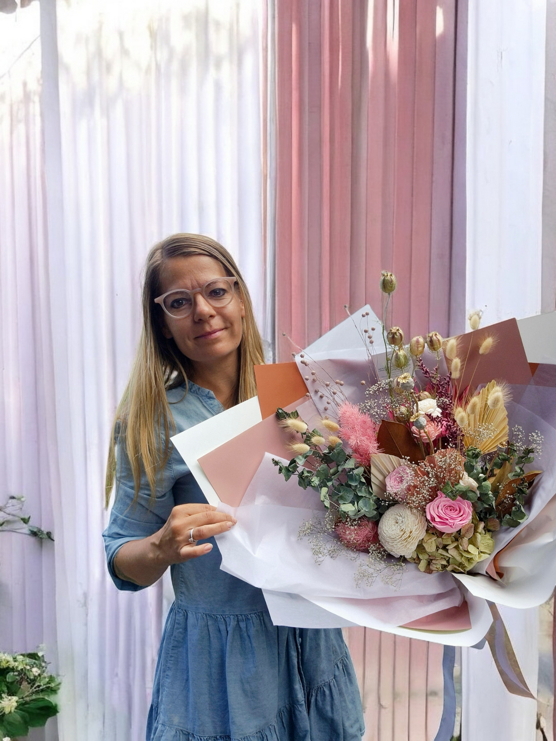 Florist Kat holds a Blush & Bloom dried floral bouquet, made with pink and white flowers and neutral foliage, wrapped in pink and white tissue paper.