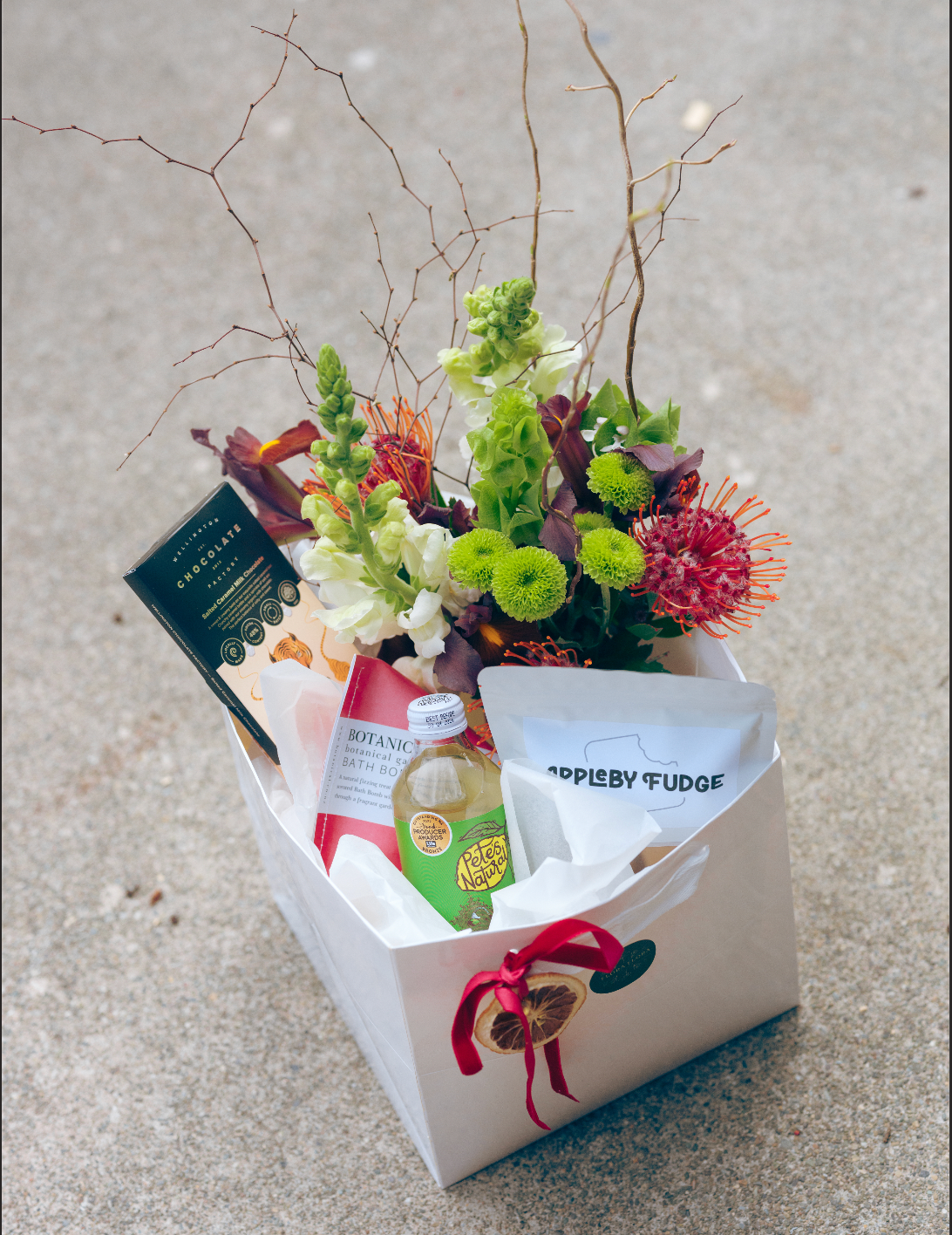 Holly Jolly Christmas gift box containing a posy of seasonal festive flowers, a glass vase, bath bomb, sparkling beverage, chocolate and fudge, presented in a white gift bag tied with festive ribbon.