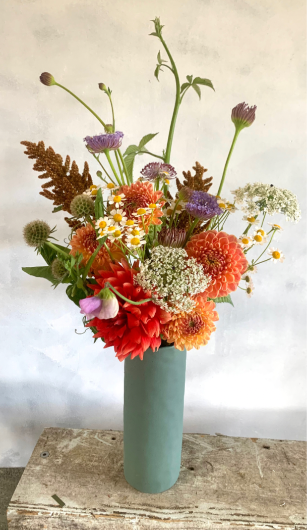 Glow floral arrangement made with orange, red, peach and lilac coloured flowers, presented in a tall green ceramic vase.