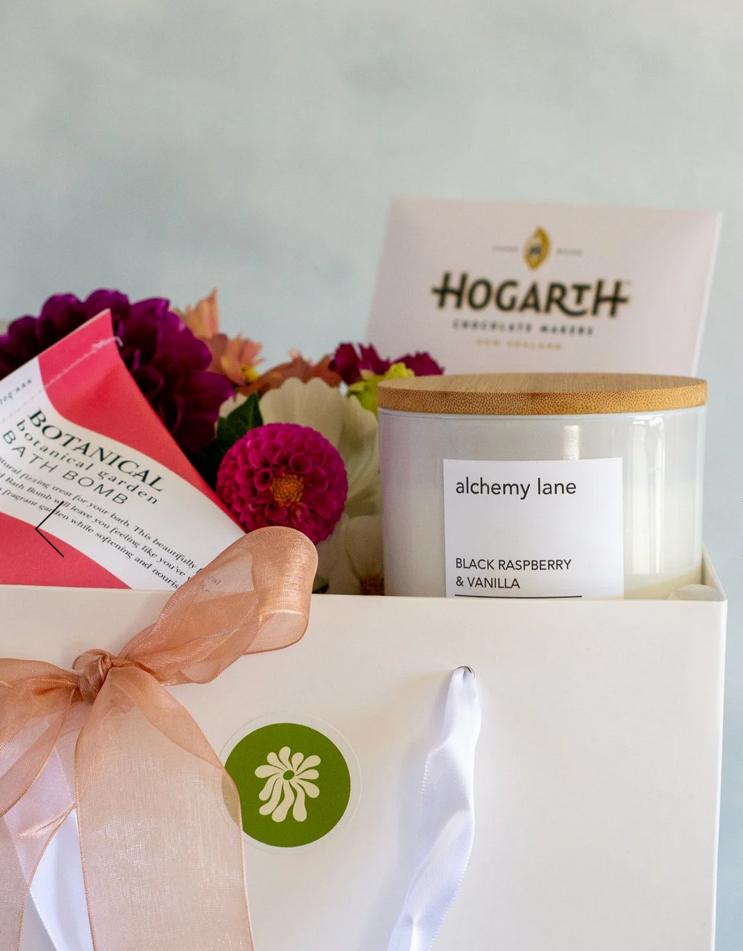 Seasonal Posie Gift Box complete with posie of fresh seasonal flowers, Botanical Bath Bomb, bar of Hogarth Chocolate, and candle by Alchemy Lane, presented in a white gift bag tied with matching ribbon.