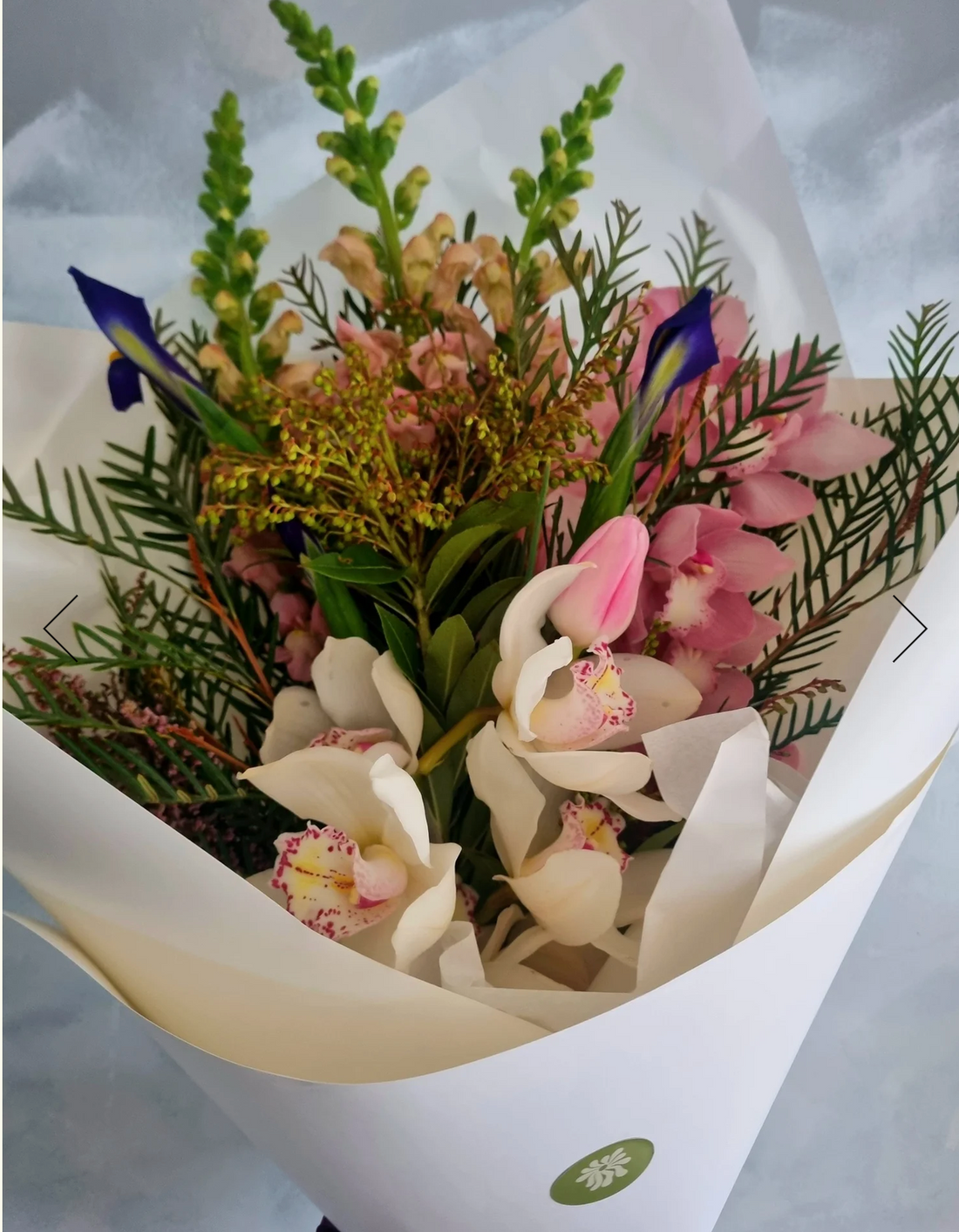 A floral bouquet with pink orchid bloom and mixed seasonal flowers and foliage in pink, white and purple, wrapped n white paper.