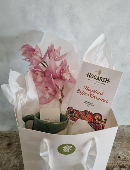 Orchid Giftbox with a beautiful pink orchid bloom, bar of Hogarth Chocolate in Hazelnut Coffee Caramel, and a handmade green mug by Francis Pottery, presented in a white gift bag.