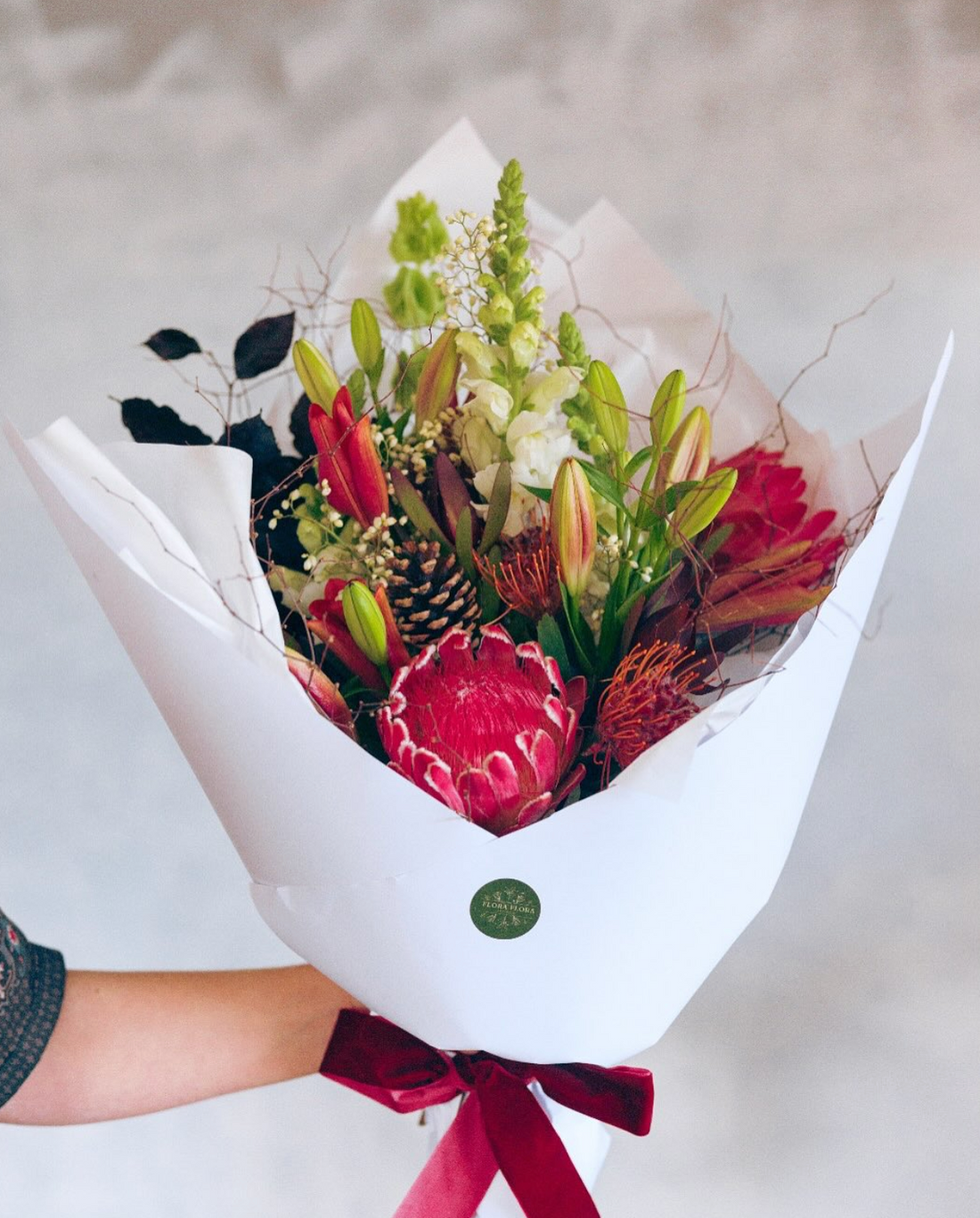 Flora Christmas bouquet made with red, orange and white flowers, wrapped in white paper and tied with a festive red ribbon.