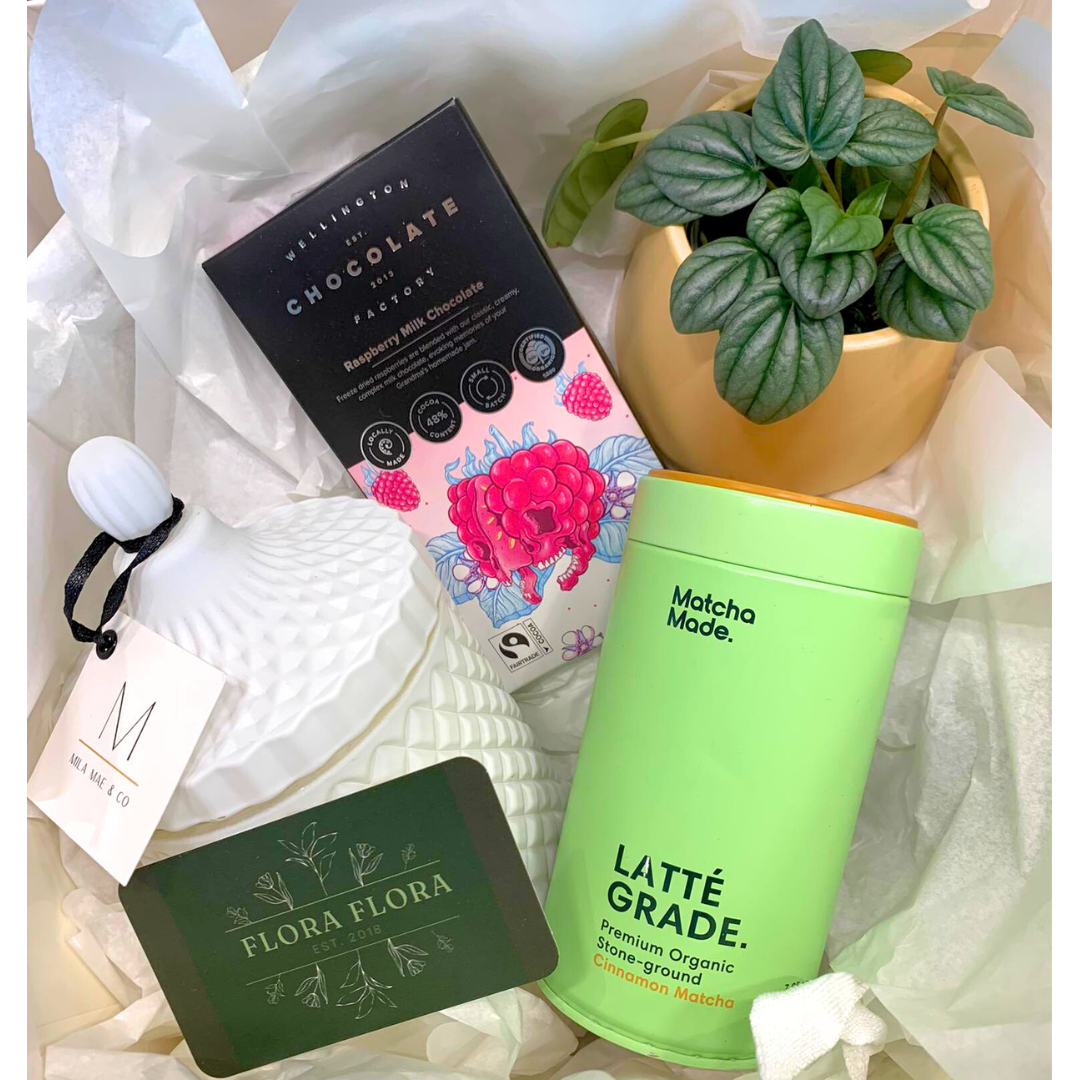 The Ultimate Gift Box containing a Mila Mae Lotus Flower Candle, Matcha Made Cinnamon Matcha, Wellington Chocolate Co bar, and a potted pepperomia moonlight plant, arranged in a box with white tissue paper.