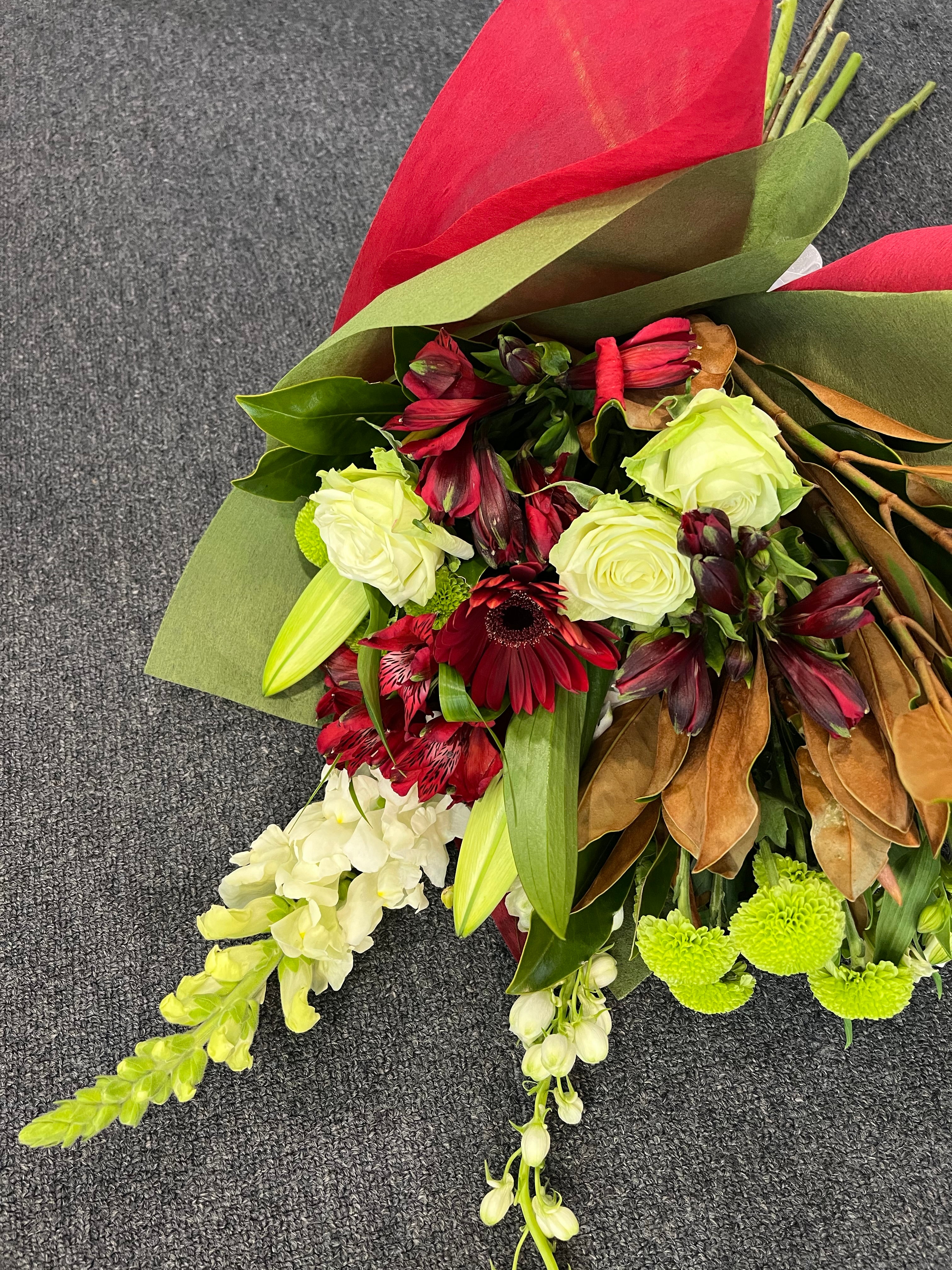 A bouquet of festive red, white and green flowers and foliage, wrapped in matching tissue paper tied with ribbon.