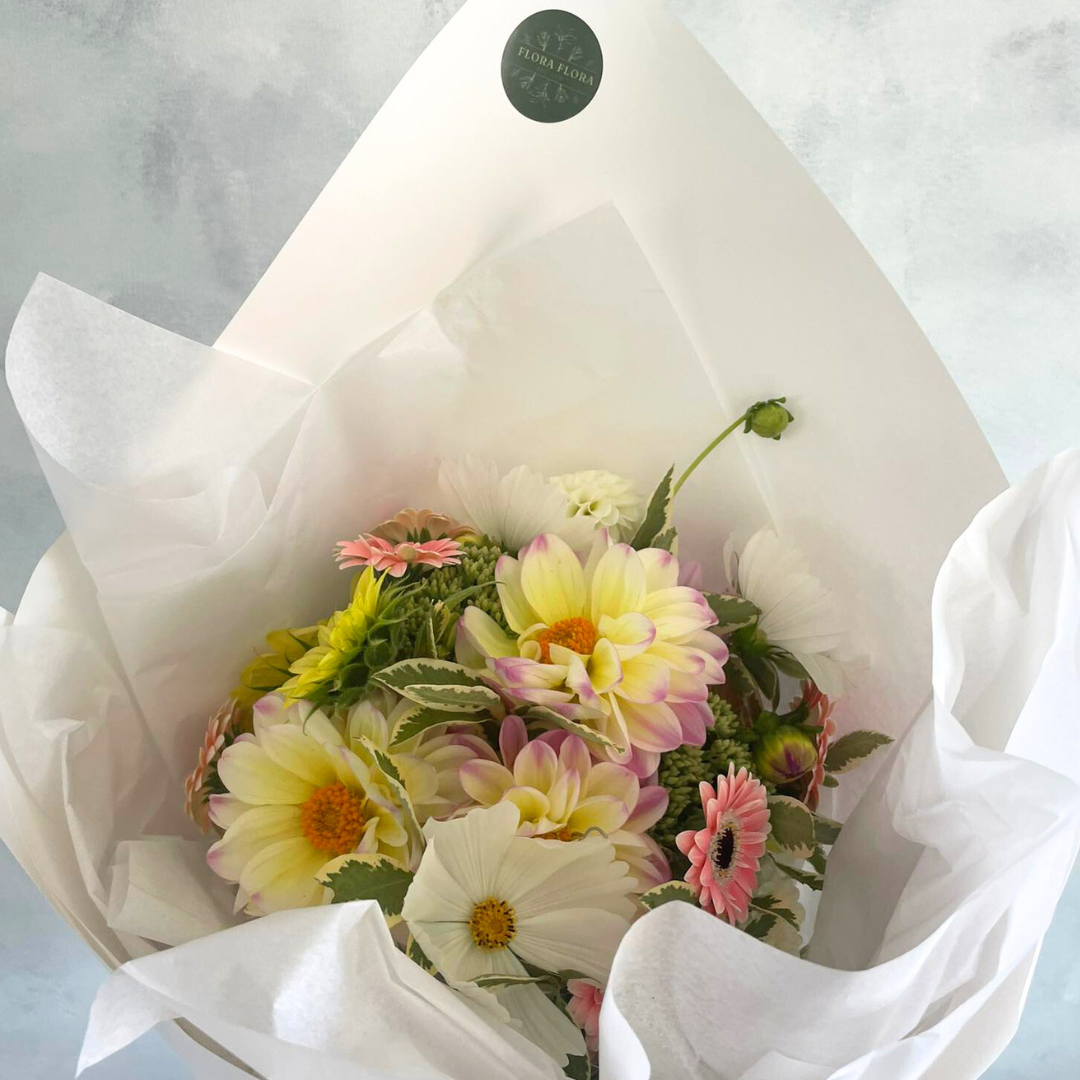 Pretty Pastels floral bouquet with soft yellow and pink flowers with greenery, wrapped in white tissue paper.